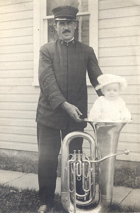 Man wearing bank uniform holding a tuba with a baby in the horn